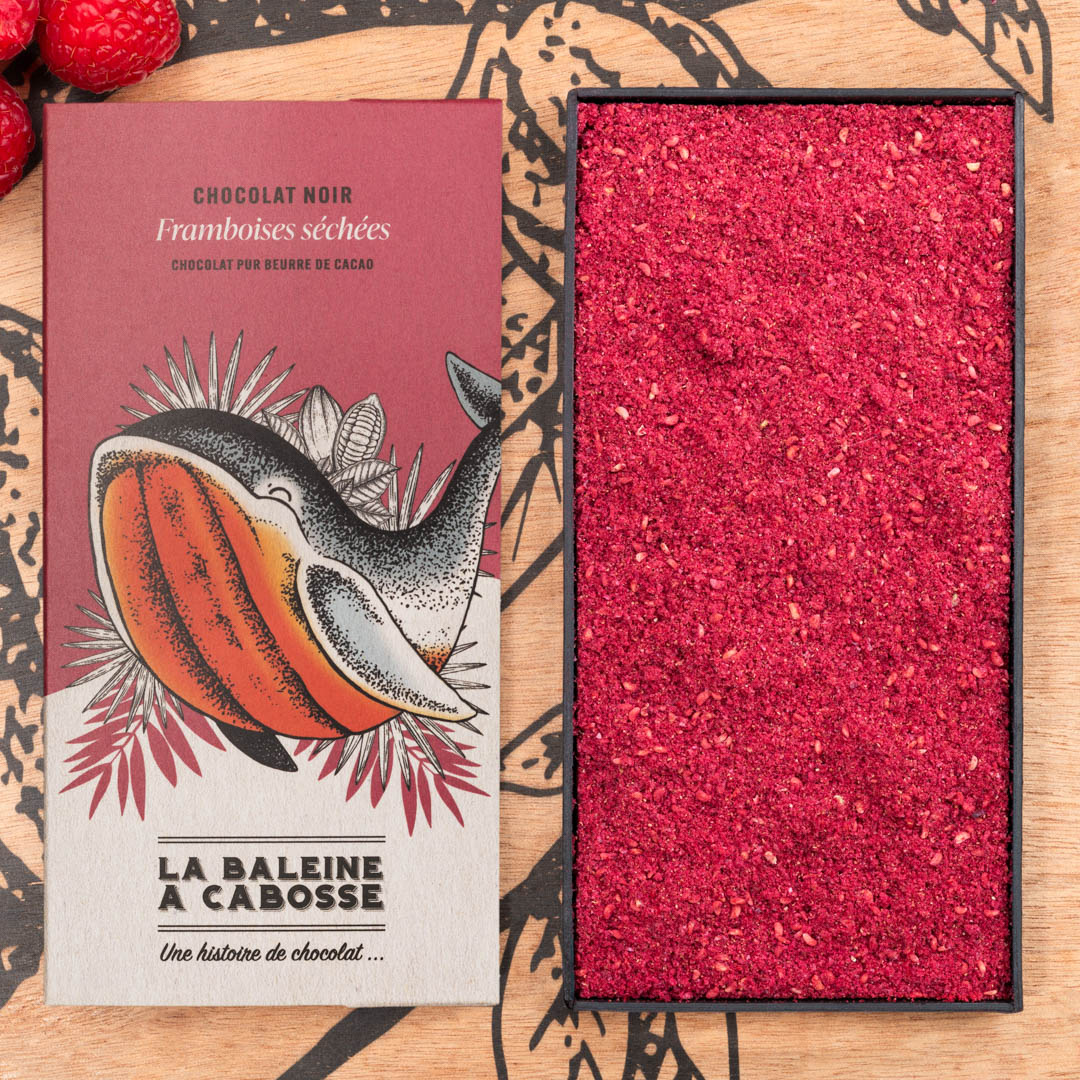 Tablette de chocolat noir 70% & framboise séchée 1 Tablette de chocolat noir 70% & framboise séchée de La Baleine à Cabosse dans son emballage, posée à côté d'un moule en forme de tablette rempli de framboise séchée en poudre