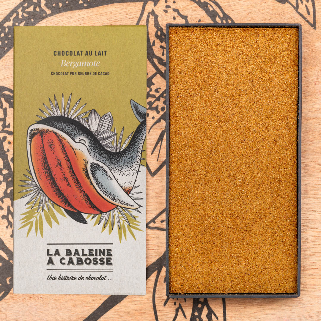 Tablette chocolat au lait 50% de cacao & bergamote 1 Tablette chocolat au lait 50% de cacao & bergamote de La Baleine à Cabosse dans son emballage posée à côté d'un moule en forme de tablette rempli de bergamote râpée