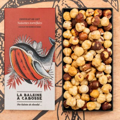 Tablette de chocolat au lait 50% de cacao & noisettes torréfiées de La Baleine à Cabosse dans son emballage, posée à côté d'un moule en forme de tablette rempli de noisettes entières
