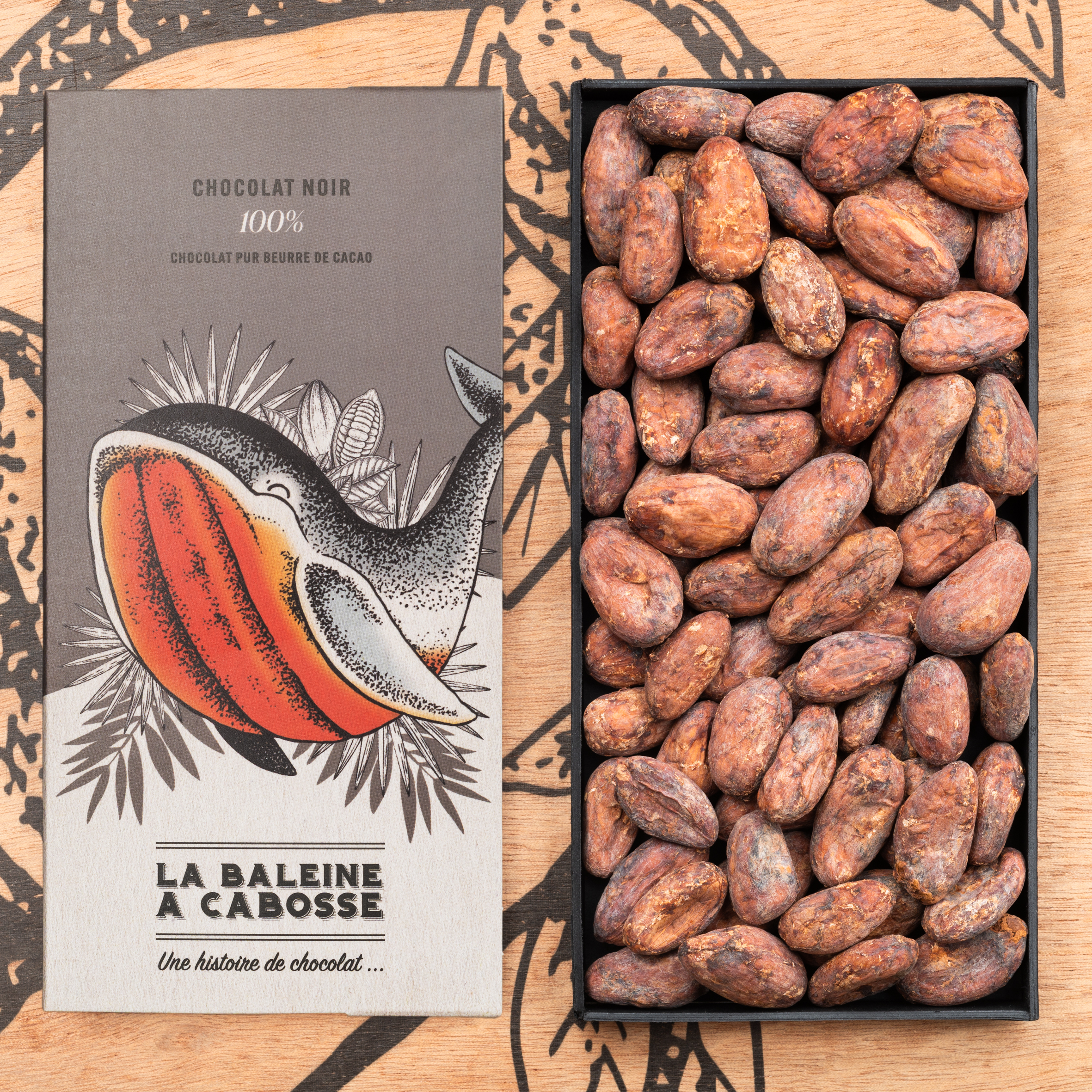 Tablette 100% Cacao 1 la tablette 100% cacao de La Baleine à Cabosse dans son emballage, posée à côté d’un moule en forme de tablette rempli de fèves torréfiées