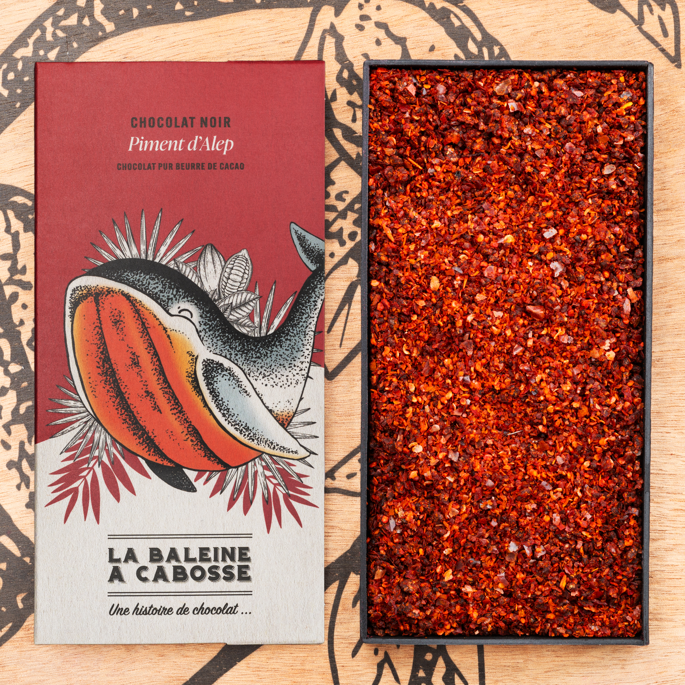 Tablette de chocolat noir 76% & piment d'Alep 1 Tablette de chocolat noir 76% & piment d'Alep de La Baleine à Cabosse dans son emballage, posée à côté d'un moule en forme de tablette rempli de piment d'Alep séché
