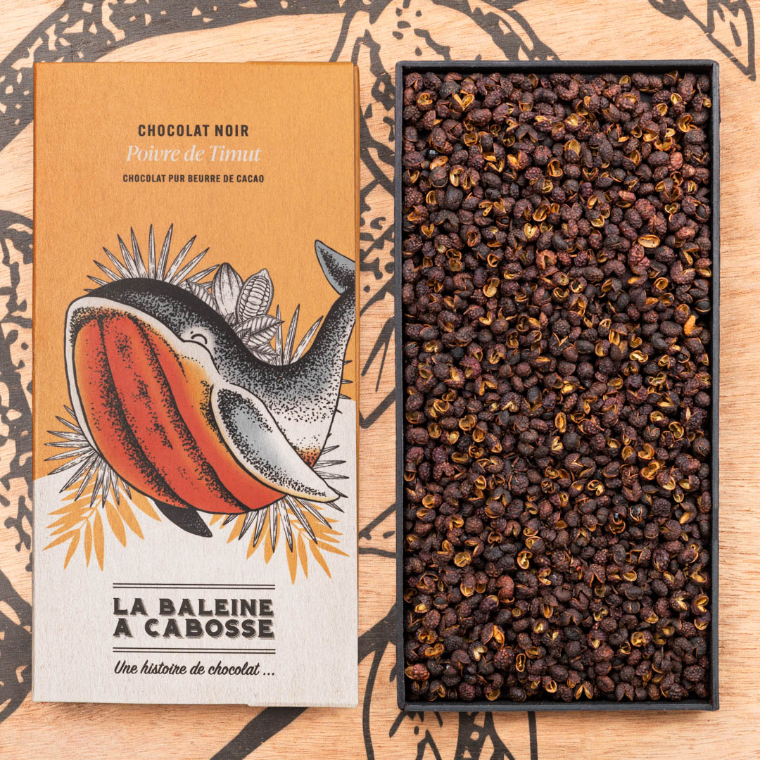 Tablette de chocolat noir 76% & poivre de timut 1 Tablette de chocolat noir 76% & poivre de timut de La Baleine à Cabosse dans son emballage, posée à côté d'un moule en forme de tablette rempli de grains de poivre de timut