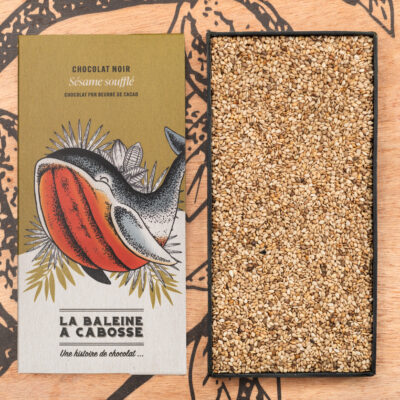 Tablette de chocolat noir 76 % & sésame soufflé de La Baleine à Cabosse dans son emballage, posée à côté d'un moule en forme de tablette rempli de grains de sésame