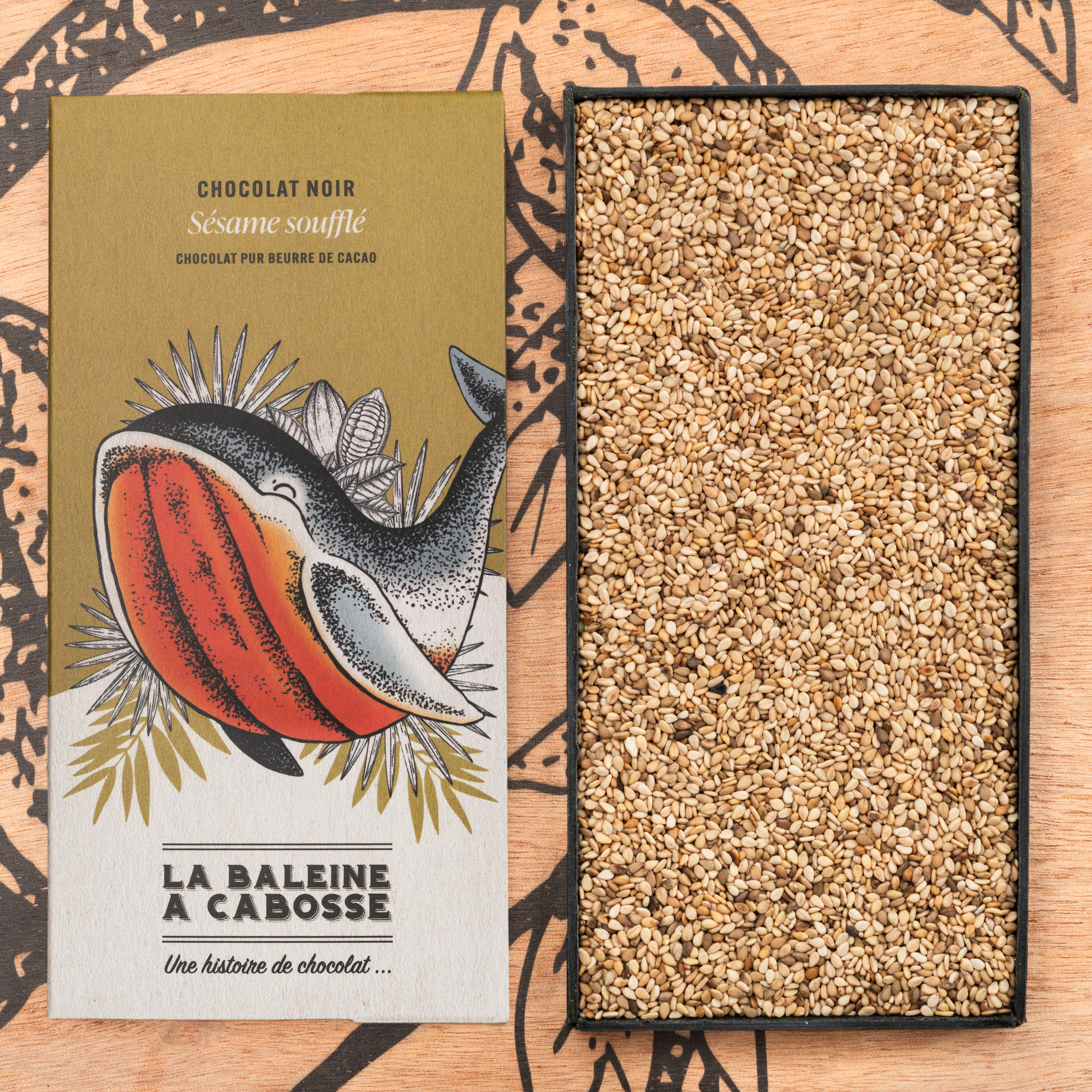 Tablette de chocolat noir 76 % & sésame soufflé 1 Tablette de chocolat noir 76 % & sésame soufflé de La Baleine à Cabosse dans son emballage, posée à côté d'un moule en forme de tablette rempli de grains de sésame