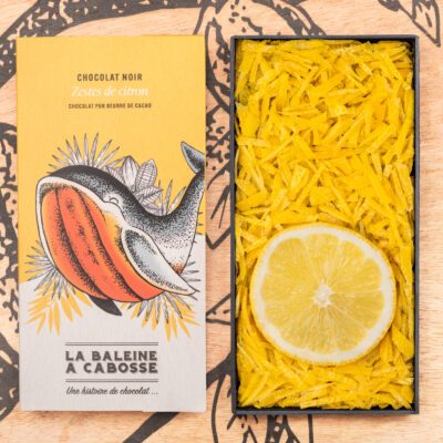 la tablette de chocolat noir aux zestes de citrons de La Baleine à Cabosse dans son emballage, posée à côté d’un moule en forme de tablette rempli de zestes de citron avec une rondelle de citron au milieu