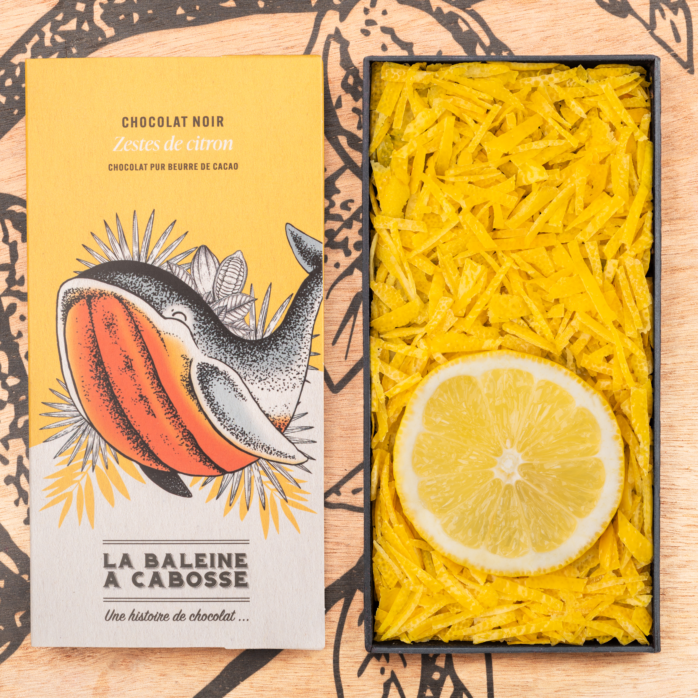 Tablette de chocolat noir 76% & zestes de citron 1 la tablette de chocolat noir aux zestes de citrons de La Baleine à Cabosse dans son emballage, posée à côté d’un moule en forme de tablette rempli de zestes de citron avec une rondelle de citron au milieu