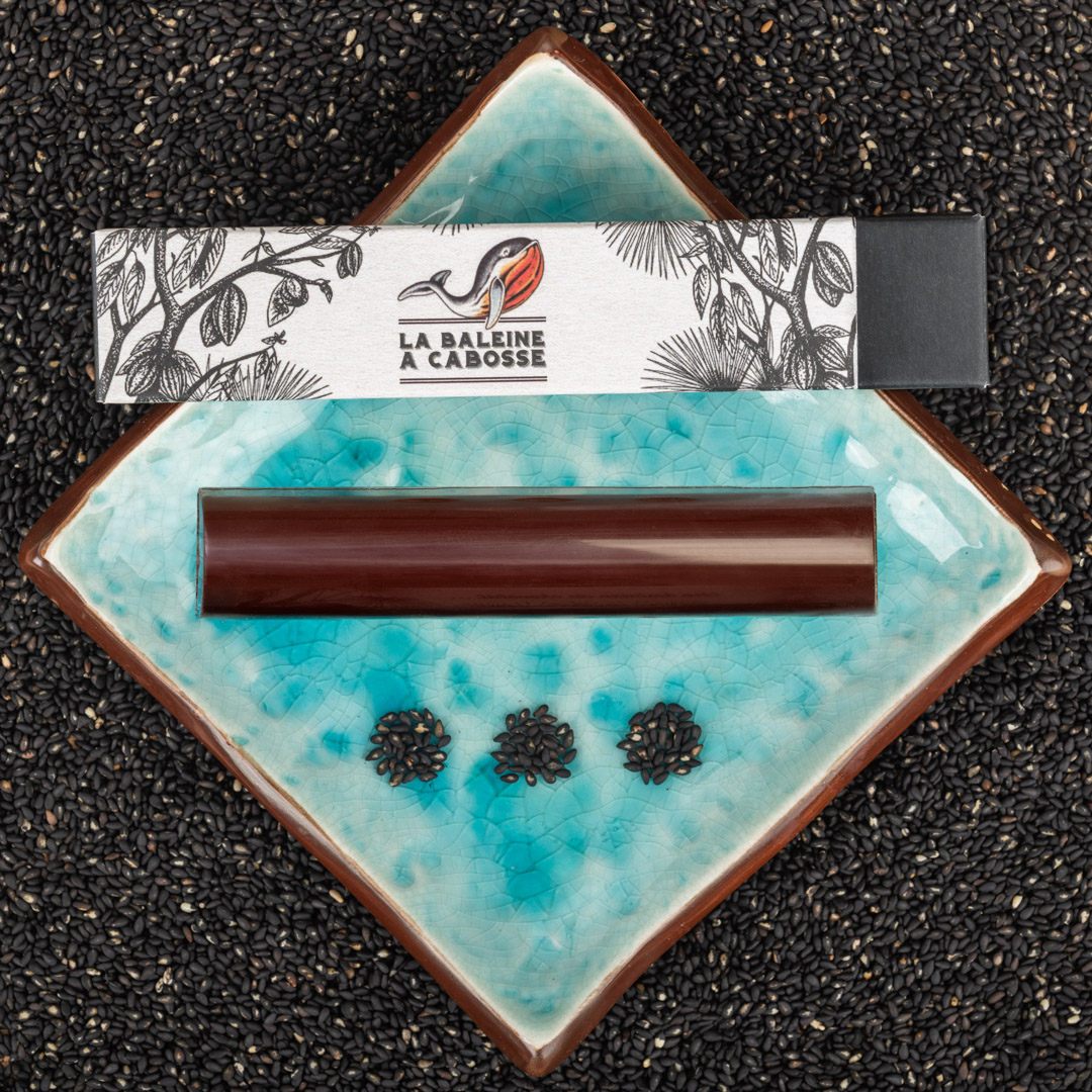 Barre fourrée au praliné de sésame noir posée à côté de son emballage sur une assiette bleue avec du sésame noir tout autour