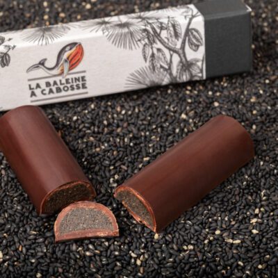 La barre fourrée au praliné de sésame noir coupée en deux et posée à côté de son emballage, sur une couche de sésame noir